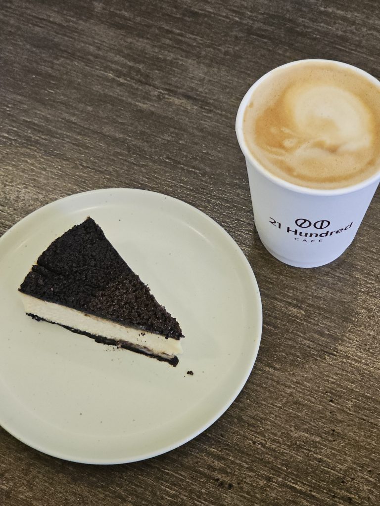 A slice of Oreo cheesecake served with a latte in a branded paper cup from 21 Hundred Cafe.